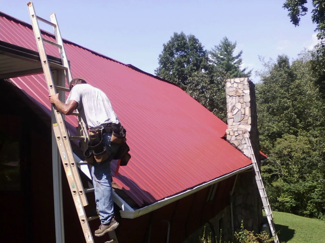 Roofer on a ladder beside a red metal roof and stone chimney — Soffit & Fascia Repair by Roofing Brothers in Lisbon